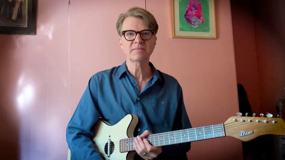 Nels Cline in a blue shirt, seated in front of pink wall with a small framed artwork, holds a worn electric guitar and looks into the camera with an announcement.